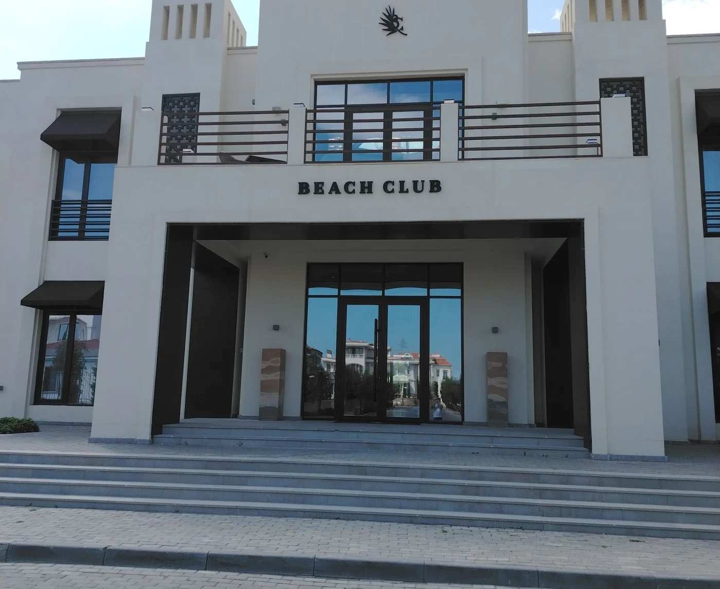 Белое здание Beach Club с надписью BEACH CLUB над входом — вход в пляжный клуб, рядом с которым стоит кофейня