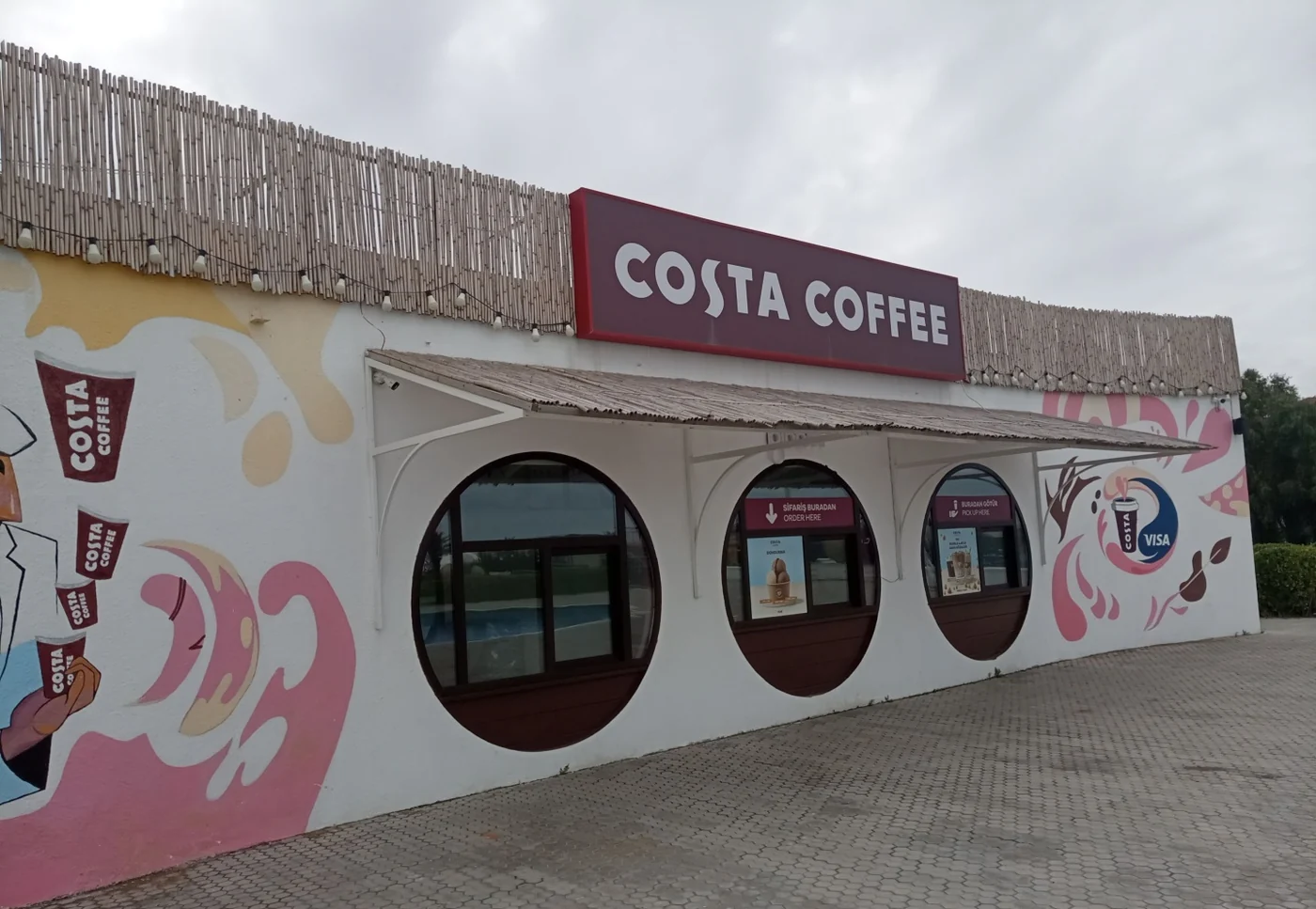 Павильон Costa Coffee в Sea Breeze прямо: три круглые арки в белой стене, тростниковый навес, бордовая вывеска и роспись с чашками по бокам