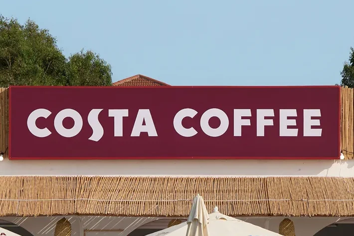 Бордовая вывеска COSTA COFFEE над тростниковым забором, слева — расписанная стена — фрагмент того же кадра